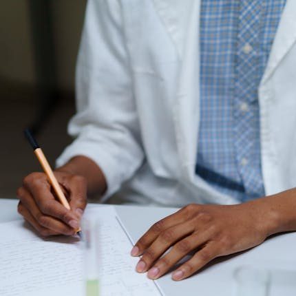 A student in a lab coat writes notes beside flasks filled with colored liquids in a laboratory setting.